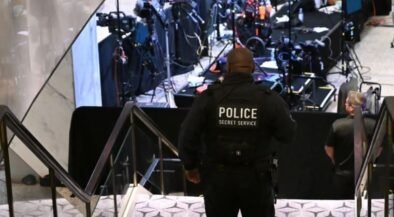 Police officer with 'POLICE SECRET SERVICE' on back stands at bottom of stairs overseeing a media briefing area with cameras and equipment in the background