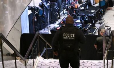 Police officer with 'POLICE SECRET SERVICE' on back stands at bottom of stairs overseeing a media briefing area with cameras and equipment in the background