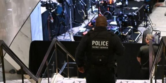Police officer with 'POLICE SECRET SERVICE' on back stands at bottom of stairs overseeing a media briefing area with cameras and equipment in the background