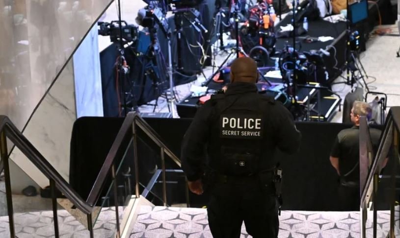 Police officer with 'POLICE SECRET SERVICE' on back stands at bottom of stairs overseeing a media briefing area with cameras and equipment in the background