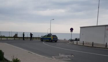 Police car blocks a street by the seaside as officers talk near a no-parking sign on the promenade edge of the road.