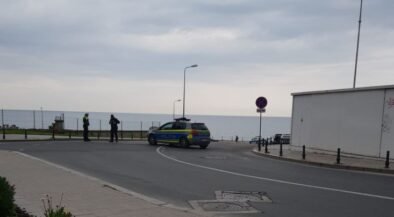Police car blocks a street by the seaside as officers talk near a no-parking sign on the promenade edge of the road.