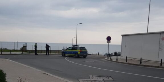 Police car blocks a street by the seaside as officers talk near a no-parking sign on the promenade edge of the road.