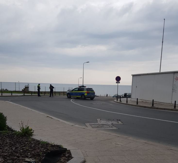 Police car blocks a street by the seaside as officers talk near a no-parking sign on the promenade edge of the road.