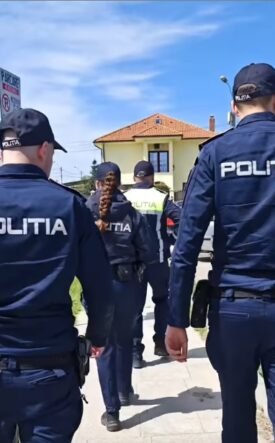 Group of police officers in dark blue uniforms labeled POLITIA walking along a sidewalk past a yellow house.