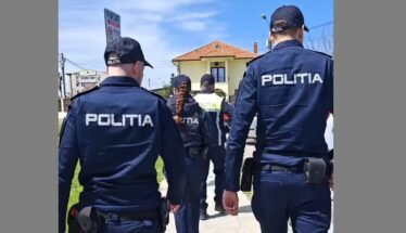 Group of police officers in dark blue uniforms labeled POLITIA walking along a sidewalk past a yellow house.