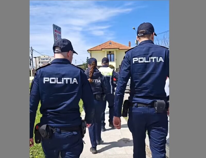 Group of police officers in dark blue uniforms labeled POLITIA walking along a sidewalk past a yellow house.