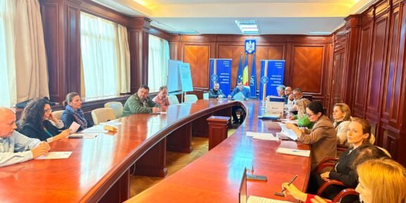 Group of professionals seated around a large curved wooden conference table in a formal meeting room, taking notes with documents and laptops nearby, banners and a crest on the back wall.
