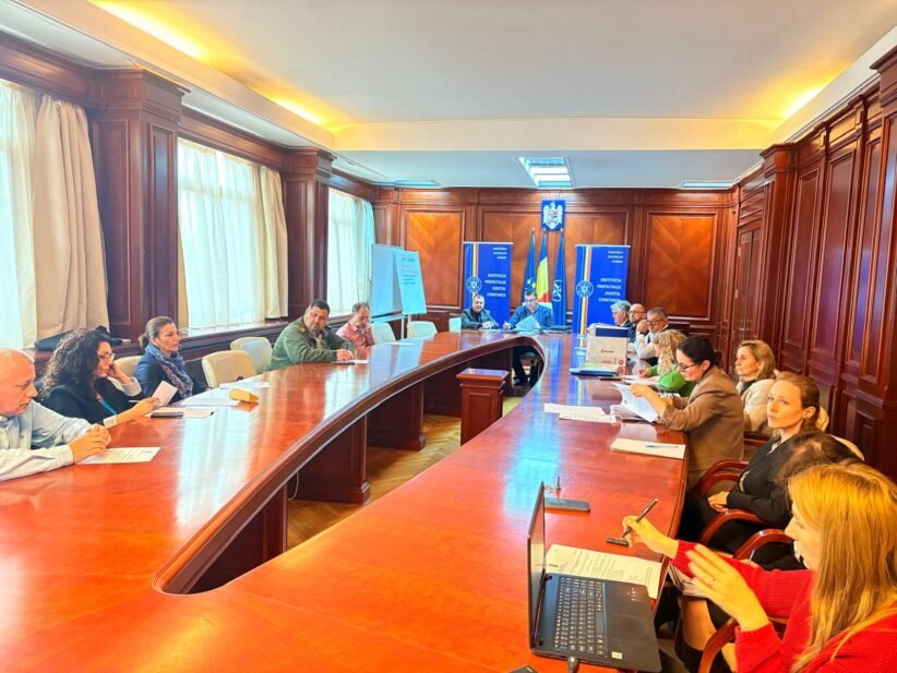 Group of professionals seated around a large curved wooden conference table in a formal meeting room, taking notes with documents and laptops nearby, banners and a crest on the back wall.