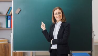 Female teacher in a black blazer pointing at a green chalkboard in a classroom.