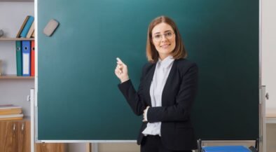 Female teacher in a black blazer pointing at a green chalkboard in a classroom.