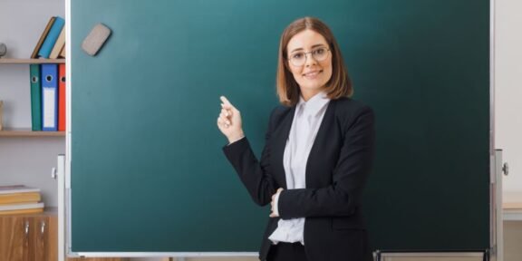 Female teacher in a black blazer pointing at a green chalkboard in a classroom.