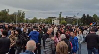 Large crowd gathered in an open square, people talking and mingling on a cloudy day with trees and buildings in the background.