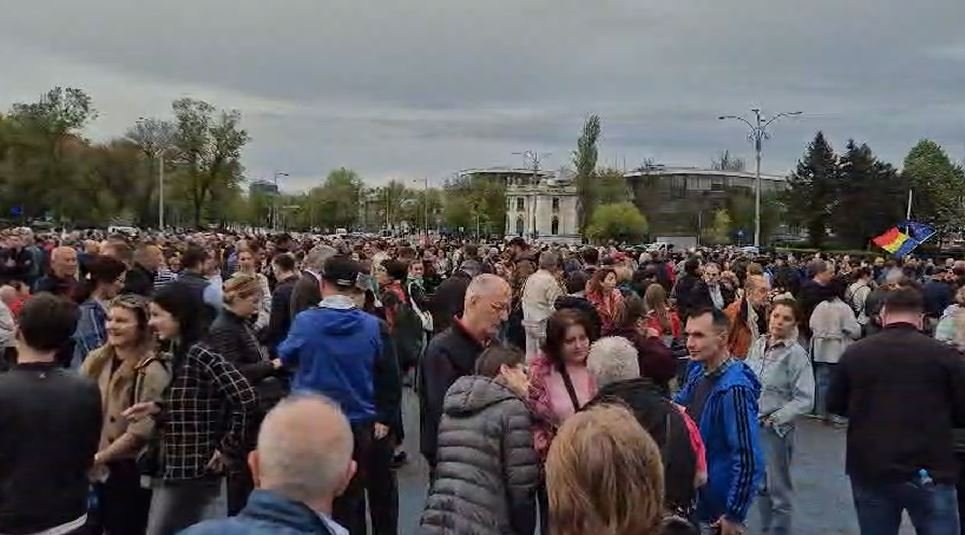 Large crowd gathered in an open square, people talking and mingling on a cloudy day with trees and buildings in the background.