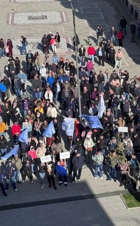 Aerial view of a large crowd in a city square participating in a protest, holding blue flags and white placards.