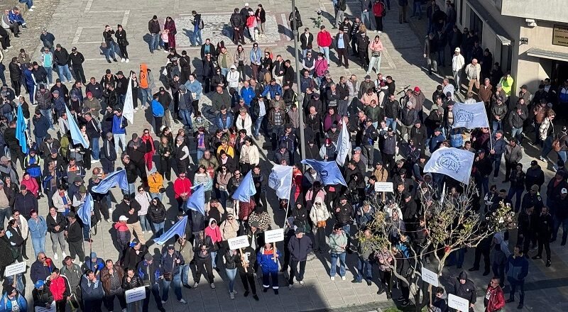Aerial view of a large crowd in a city square participating in a protest, holding blue flags and white placards.
