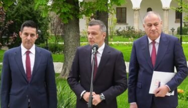 Three men in suits stand outdoors at a press conference, with a microphone in front of the center speaker.