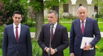 Three men in suits stand outdoors at a press conference, with a microphone in front of the center speaker.