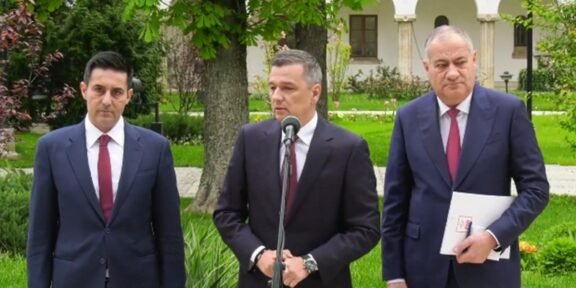 Three men in suits stand outdoors at a press conference, with a microphone in front of the center speaker.