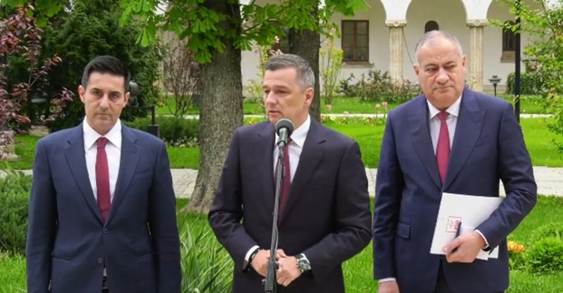 Three men in suits stand outdoors at a press conference, with a microphone in front of the center speaker.