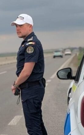Policeman in dark uniform and white cap standing beside a police SUV on a highway, looking toward oncoming traffic as stormy clouds loom.