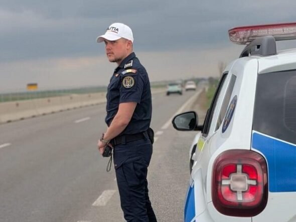 Policeman in dark uniform and white cap standing beside a police SUV on a highway, looking toward oncoming traffic as stormy clouds loom.