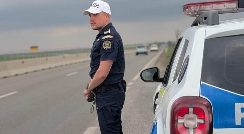 Policeman in dark uniform and white cap standing beside a police SUV on a highway, looking toward oncoming traffic as stormy clouds loom.