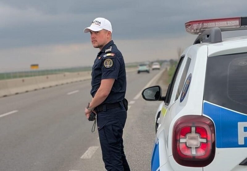 Policeman in dark uniform and white cap standing beside a police SUV on a highway, looking toward oncoming traffic as stormy clouds loom.