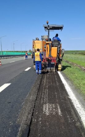 Road construction: a milling machine removing asphalt on a highway with workers in high-vis vests nearby, blue sky above.