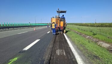 Road construction: a milling machine removing asphalt on a highway with workers in high-vis vests nearby, blue sky above.