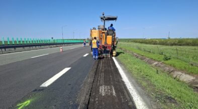 Road construction: a milling machine removing asphalt on a highway with workers in high-vis vests nearby, blue sky above.