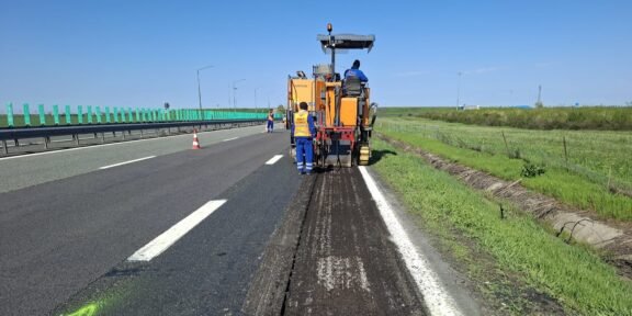 Road construction: a milling machine removing asphalt on a highway with workers in high-vis vests nearby, blue sky above.