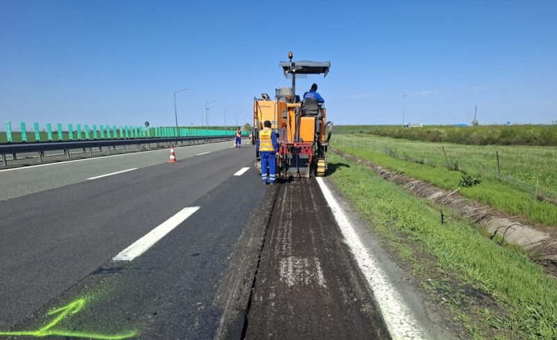 Road construction: a milling machine removing asphalt on a highway with workers in high-vis vests nearby, blue sky above.