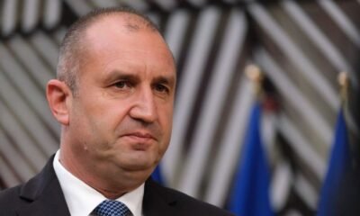 Man in a dark suit and white shirt with a blue tie, facing slightly left, at a formal event with blurred flags in the background.