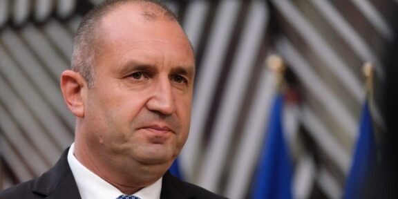 Man in a dark suit and white shirt with a blue tie, facing slightly left, at a formal event with blurred flags in the background.