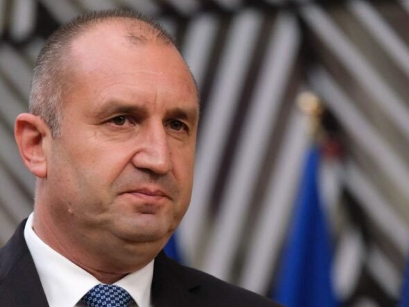 Man in a dark suit and white shirt with a blue tie, facing slightly left, at a formal event with blurred flags in the background.