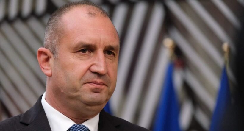 Man in a dark suit and white shirt with a blue tie, facing slightly left, at a formal event with blurred flags in the background.