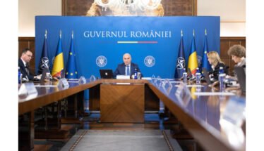 Formal government briefing in a room with a blue backdrop reading 'Guvernul României', central official at the head of a long table flanked by flags and colleagues.