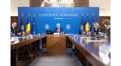 Formal government briefing in a room with a blue backdrop reading 'Guvernul României', central official at the head of a long table flanked by flags and colleagues.