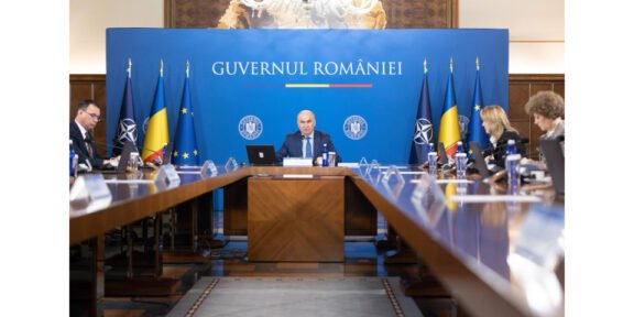 Formal government briefing in a room with a blue backdrop reading 'Guvernul României', central official at the head of a long table flanked by flags and colleagues.