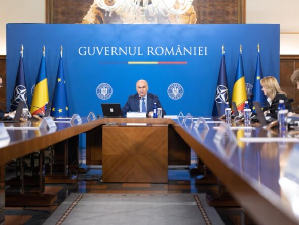 Formal government briefing in a room with a blue backdrop reading 'Guvernul României', central official at the head of a long table flanked by flags and colleagues.