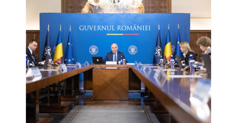 Formal government briefing in a room with a blue backdrop reading 'Guvernul României', central official at the head of a long table flanked by flags and colleagues.