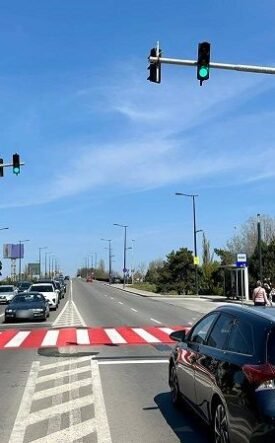Wide city street with a red-striped pedestrian crosswalk, cars waiting at a green light, and a blue sky.