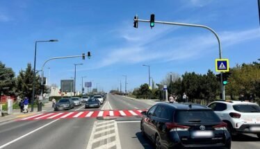 Wide city street with a red-striped pedestrian crosswalk, cars waiting at a green light, and a blue sky.