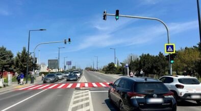 Wide city street with a red-striped pedestrian crosswalk, cars waiting at a green light, and a blue sky.