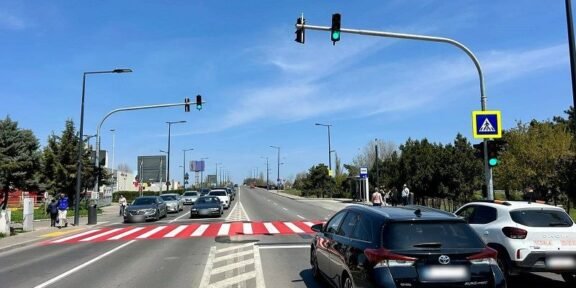 Wide city street with a red-striped pedestrian crosswalk, cars waiting at a green light, and a blue sky.