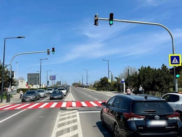 Wide city street with a red-striped pedestrian crosswalk, cars waiting at a green light, and a blue sky.