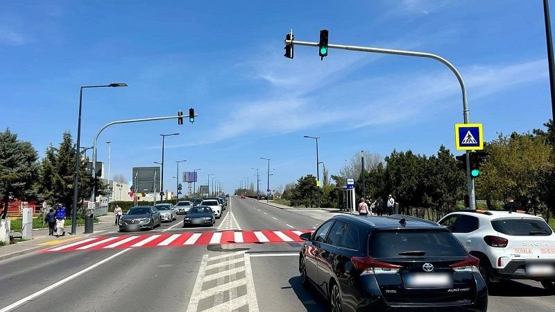 Wide city street with a red-striped pedestrian crosswalk, cars waiting at a green light, and a blue sky.