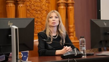 Woman with long blonde hair sits at a desk with a microphone, flanked by two computer monitors in a formal room.