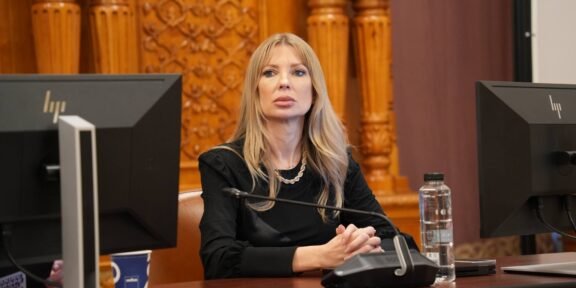 Woman with long blonde hair sits at a desk with a microphone, flanked by two computer monitors in a formal room.